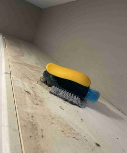 A yellow and black scrub brush with gray bristles sits on a dirty, stained countertop near a beige wall.
