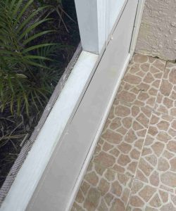 Close-up of a white screened enclosure frame next to a textured tile floor and some green plants outside.