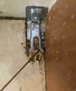 A metal bracket is mounted at the junction of a tiled floor and wall, with visible dirt and stains around it.