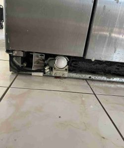 The lower section of a stainless steel appliance with the front panel removed, exposing internal components and wiring.