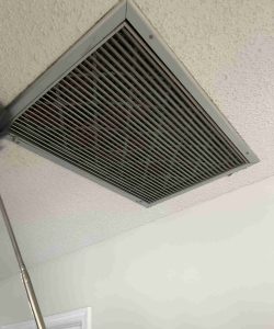 A rectangular ceiling vent with dark metal slats is installed on a textured, off-white ceiling.