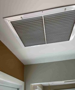 A large rectangular ceiling air vent with metal slats is installed near a window in a room with beige and white walls.