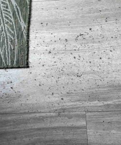 Gray tile floor with scattered dust and debris next to the corner of a green patterned rug.