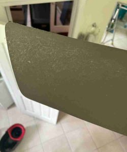 A dusty ceiling fan blade shown close up, with a tiled floor and household items visible in the background.
