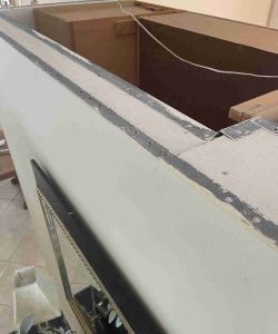 Close-up of a partially constructed interior wall with exposed edges and metal strips, overlooking wooden cabinets.