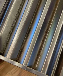 Close-up of a metal vent or filter with visible dirt buildup and rainbow-like discoloration on the surface.