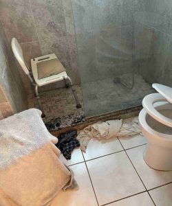 Accessible shower with a chair, towels and clothes on the floor, and an open toilet in a tiled bathroom.