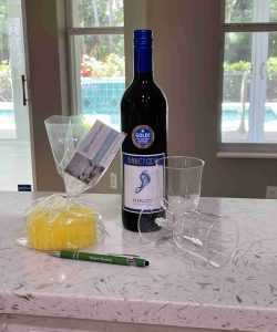 A bottle of Barefoot Merlot, two plastic wine glasses, a wrapped sponge, a note, and a green pen on a counter.