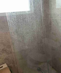 A glass shower panel covered in water droplets, with a tiled wall and a small window in the background.